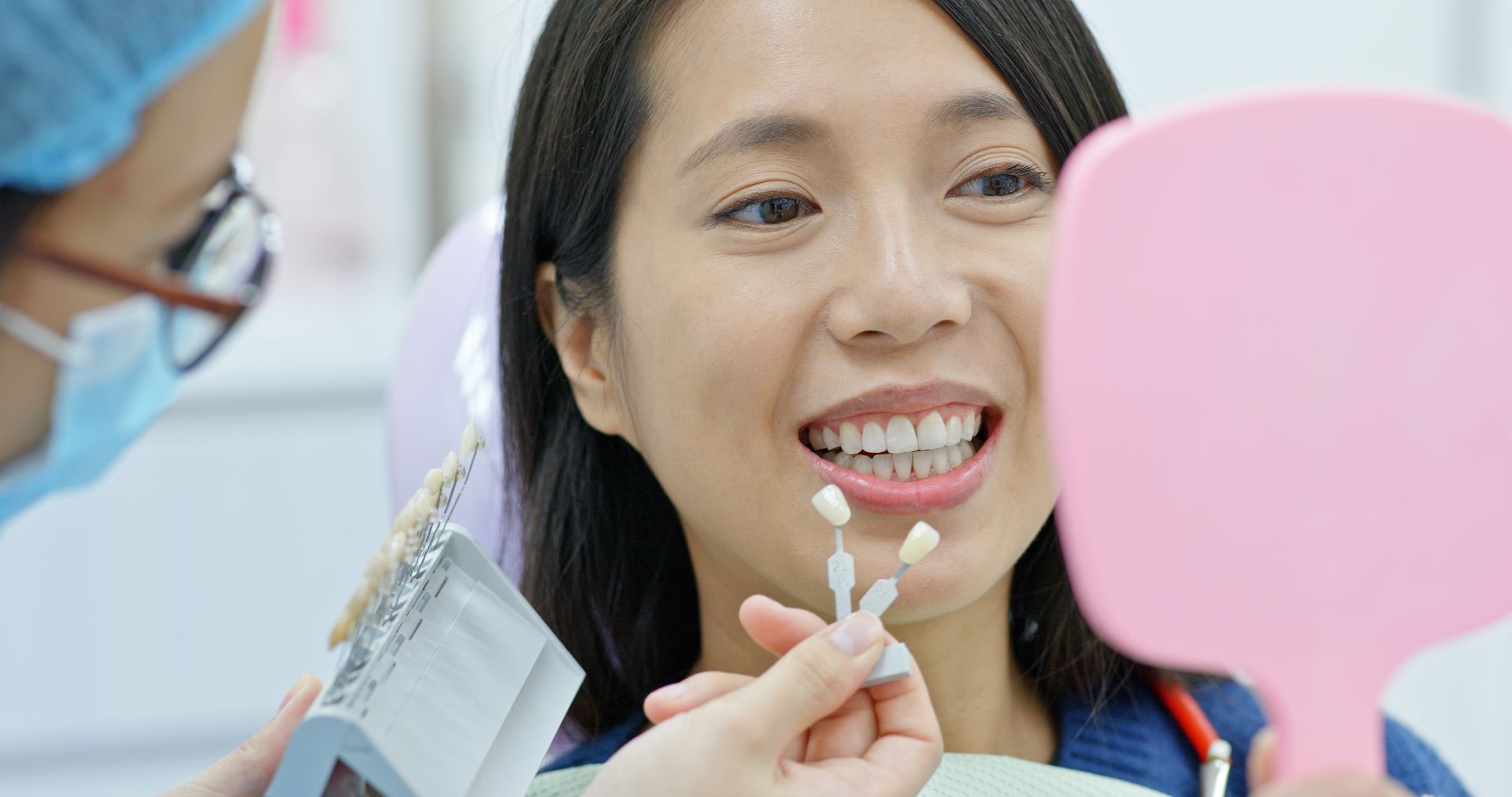 Woman at dentist with teeth shade samples, smiling; dentist holds mirror and shade guide.
