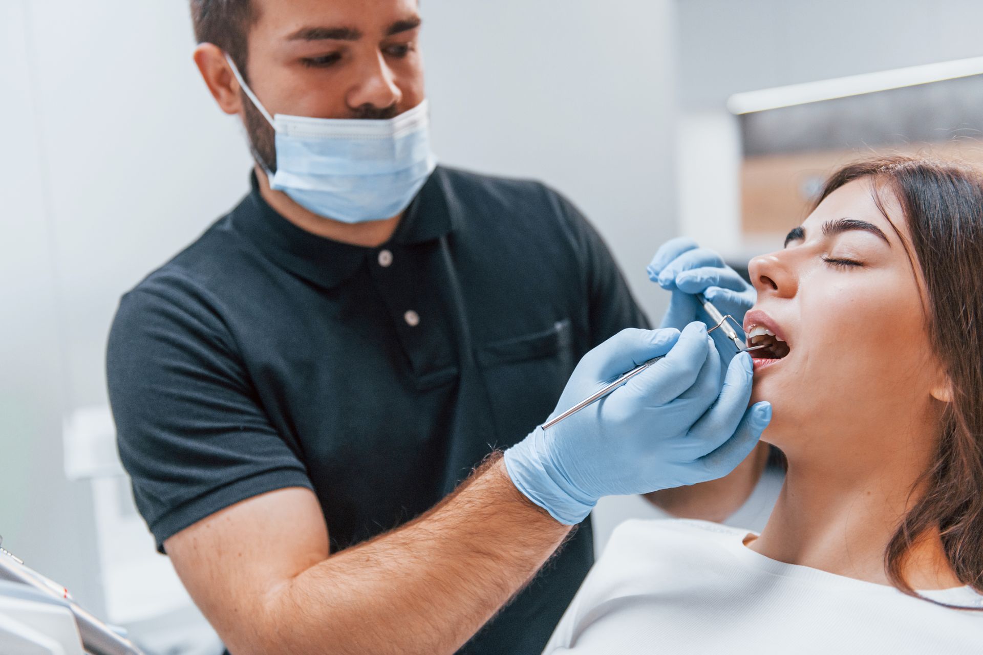 Dentist examining a woman's teeth in a clinic. The dentist wears a mask and gloves. The woman has her mouth open.