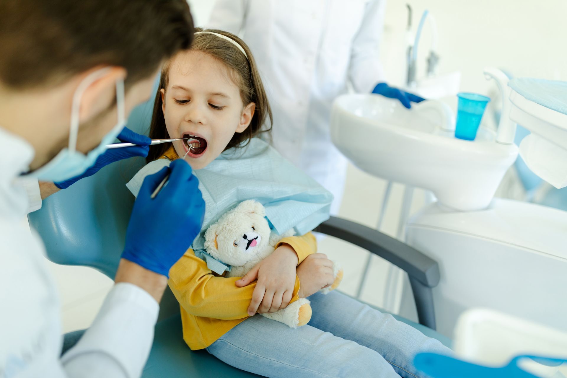 Child in dentist chair, mouth open, examined by dentist with tools. Includes teddy bear.