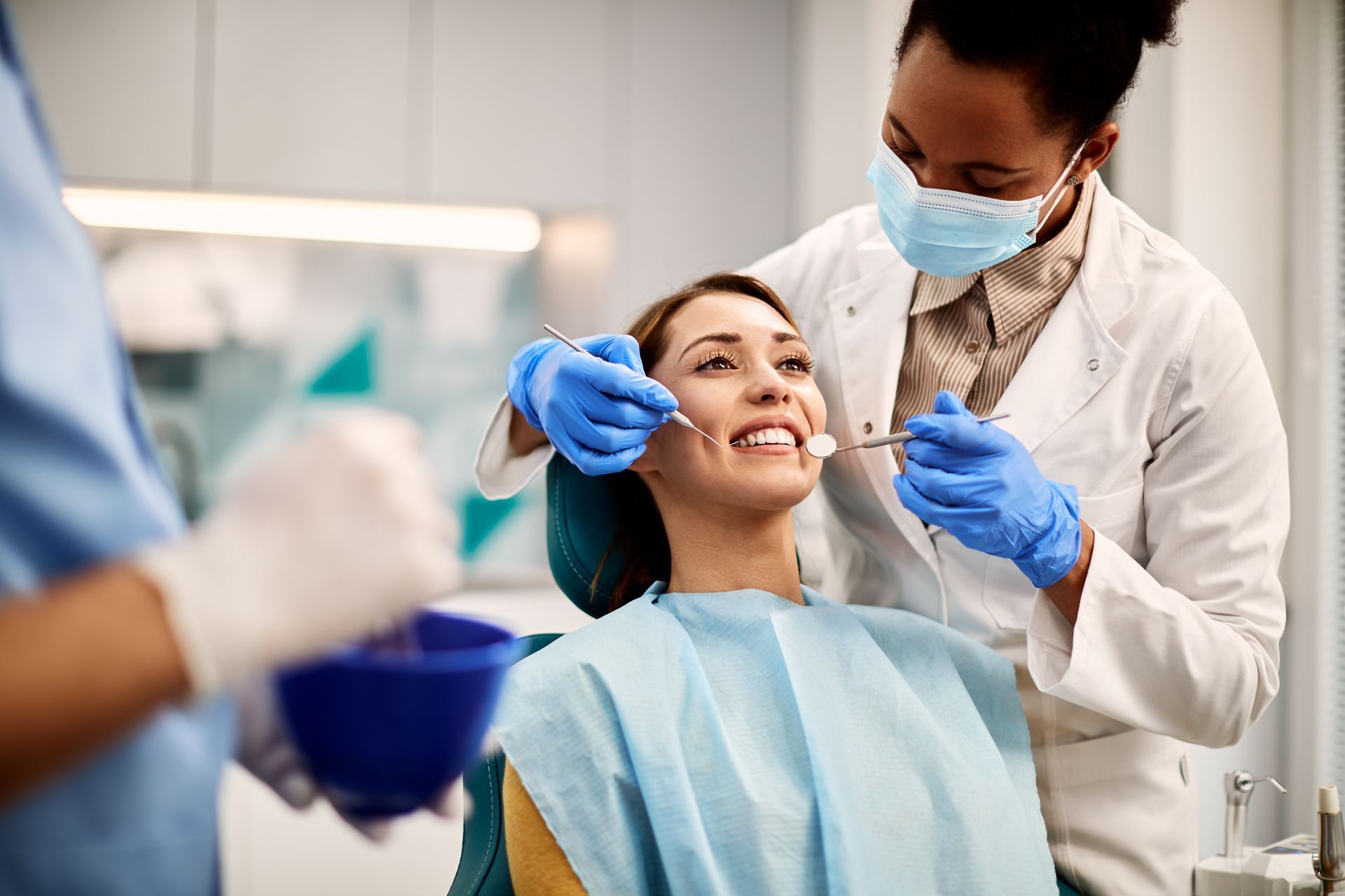 Dentist examining patient's teeth with tools in a dental office. The patient smiles, wearing a protective bib.