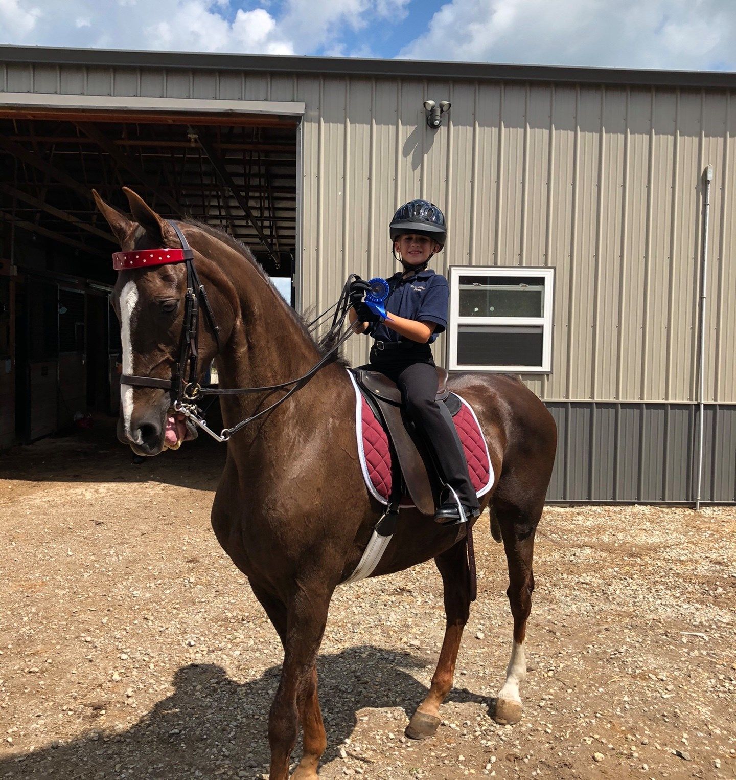 A child in riding gear on a brown horse, in front of a tan building.
