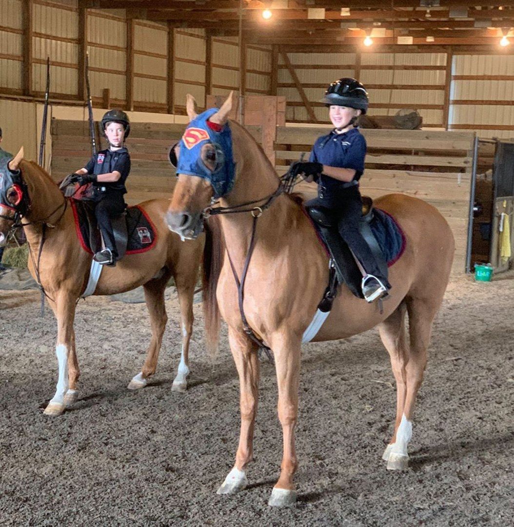 Two children on horseback in a stable; one horse wears a Superman mask.