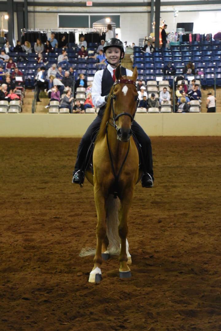 A rider on a tan horse in an indoor arena, facing forward. The rider is smiling. Spectators are in the background.
