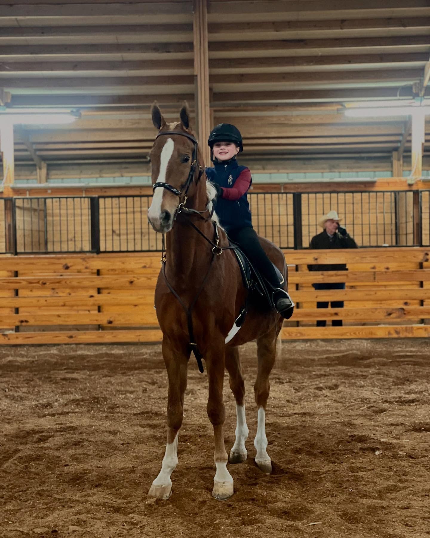 Young rider on brown horse in indoor arena, smiling.