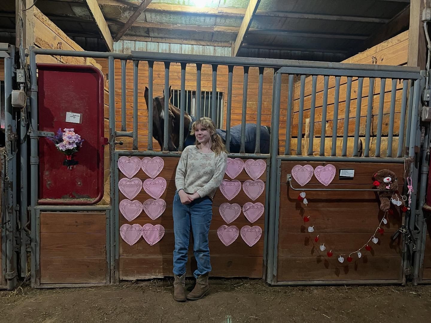 Woman in a barn leans against a stall decorated with hearts, horse in the background.