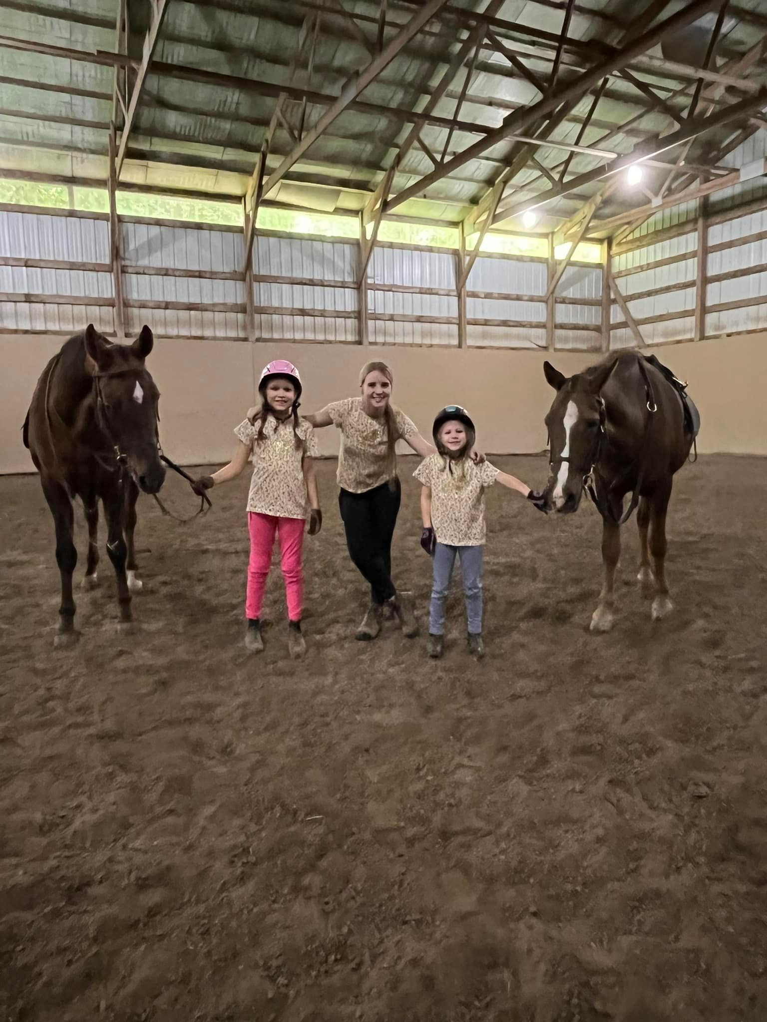 Three people with two horses inside an indoor riding arena. Two children, one adult. All smiling.