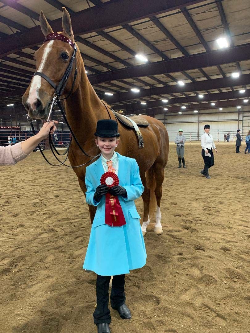 Young rider in blue coat holding a red ribbon, posing with a chestnut horse in an indoor arena.
