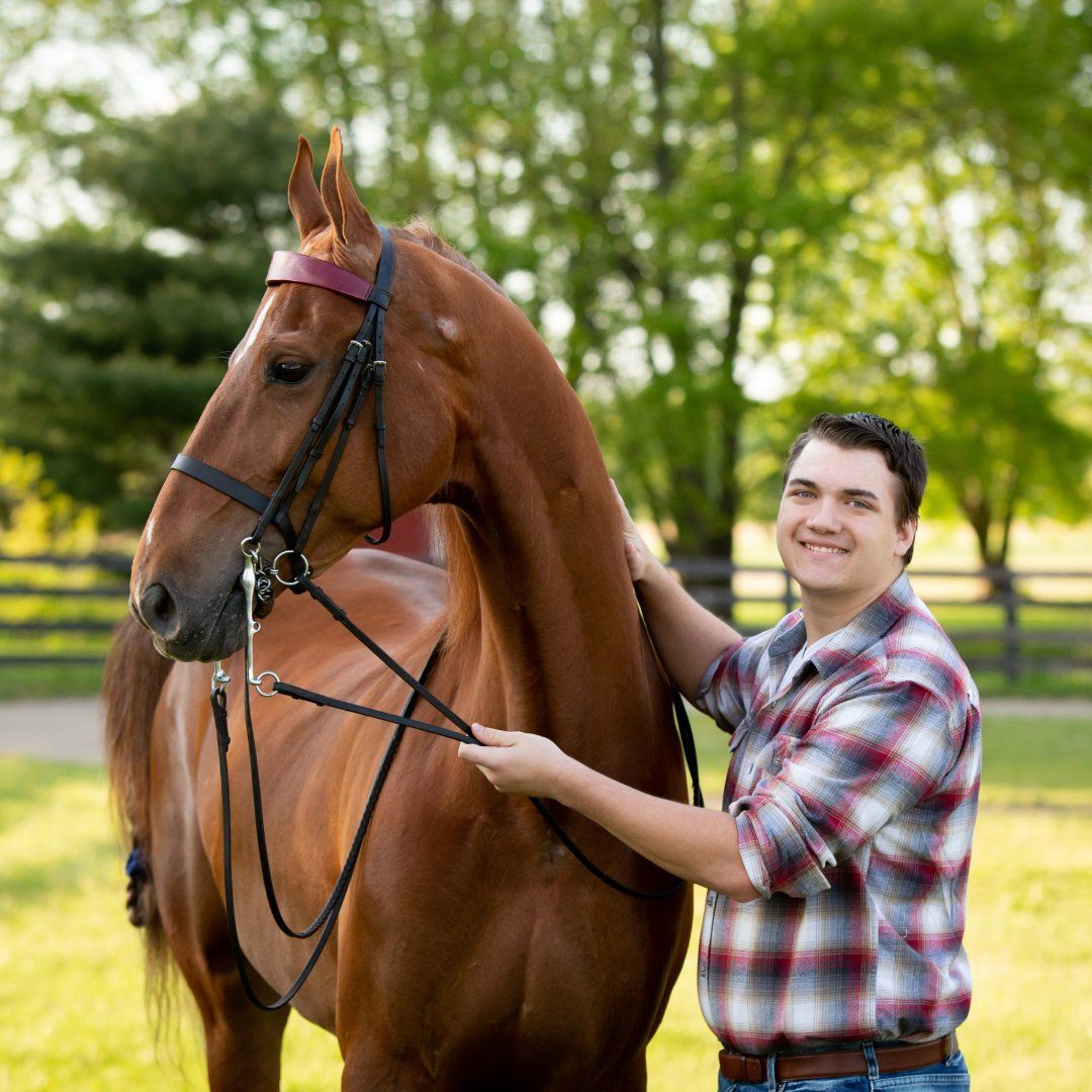 Man in plaid shirt smiles, holding reins of a brown horse. Green background, sunny outdoor setting.
