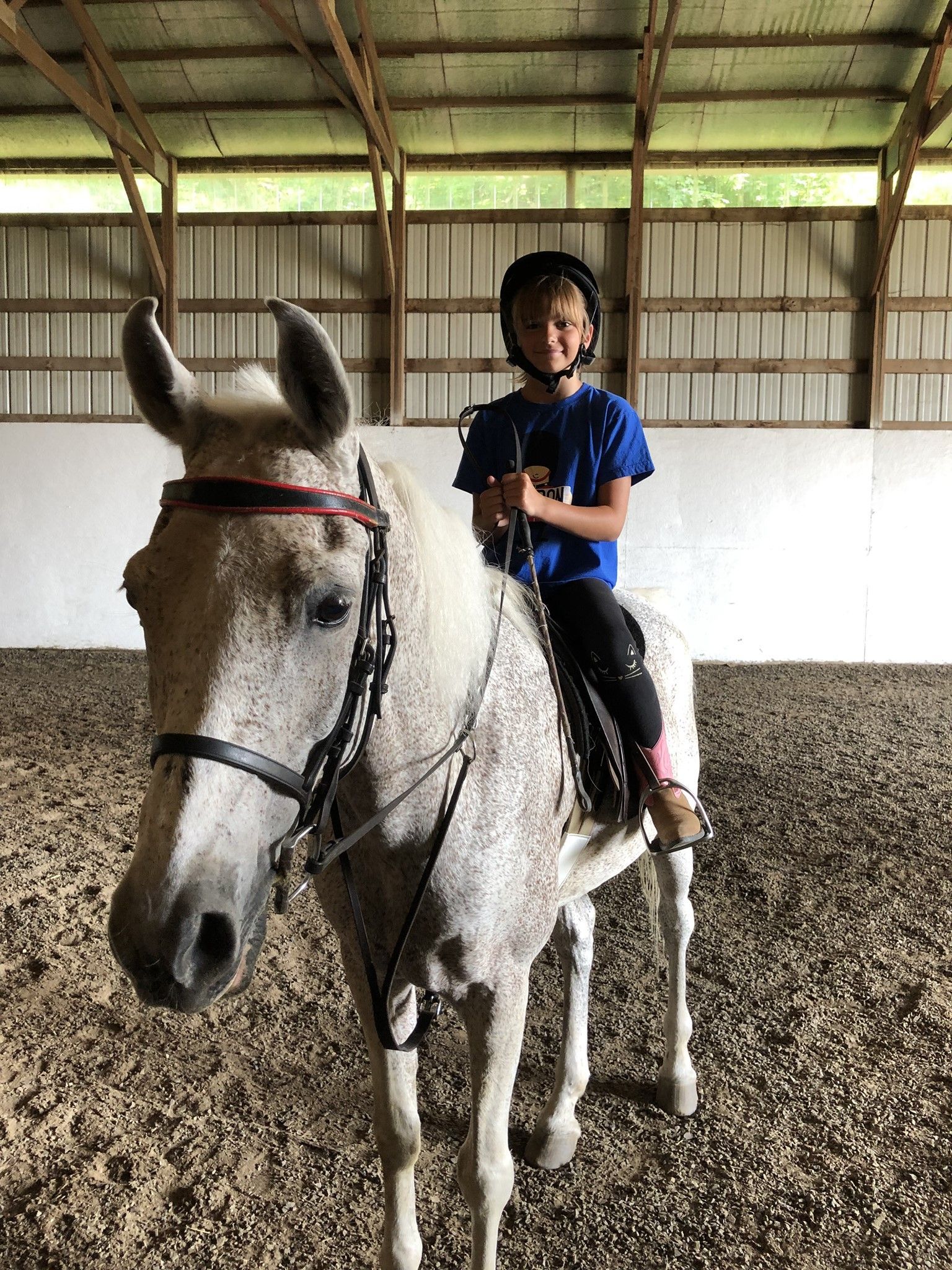 Young person on a gray horse in an indoor riding arena; wearing a helmet and blue shirt, smiling.