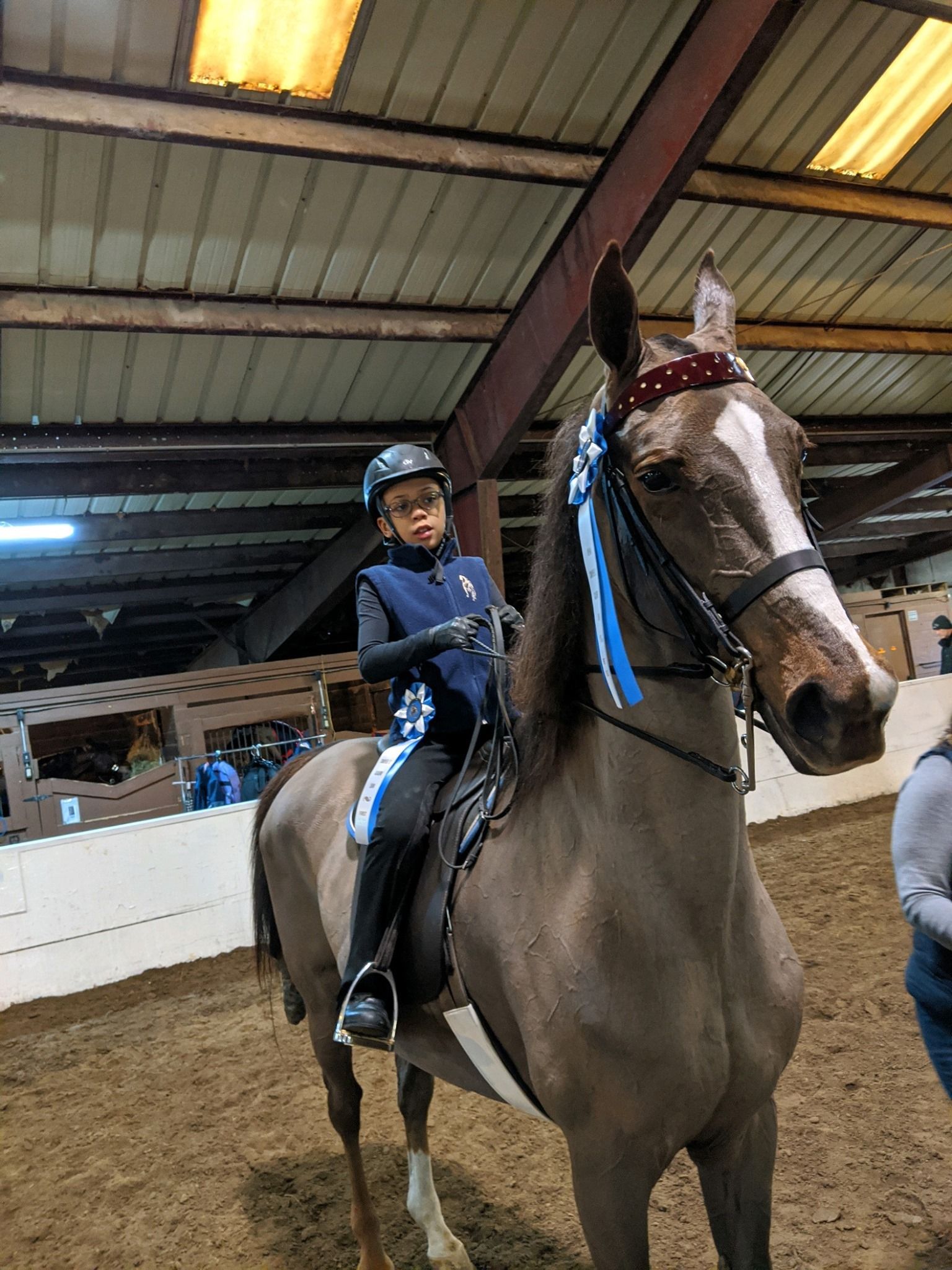 A child on a gray horse in an indoor arena, wearing a helmet and riding attire. Horse has ribbons on its bridle.