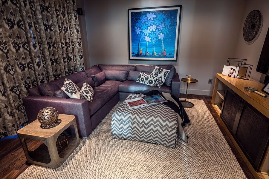 A living room with a purple couch , ottoman , table and painting on the wall.