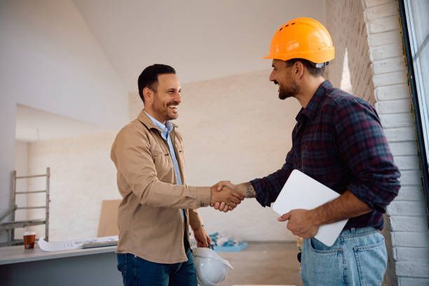 Two men shaking hands in a construction site; one in a hard hat, smiling.