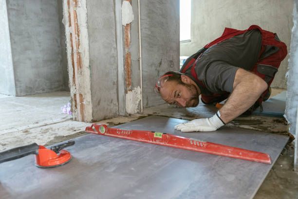 A person installing floor tiles, using a level. Construction setting, gray tiles, and concrete walls.