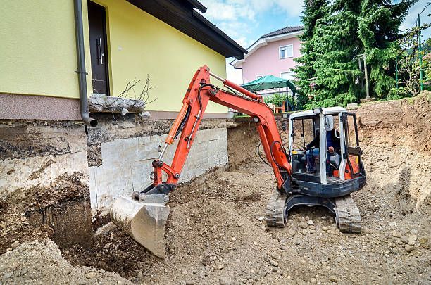 A small excavator with a red arm digs beside a house's foundation, surrounded by mounds of earth