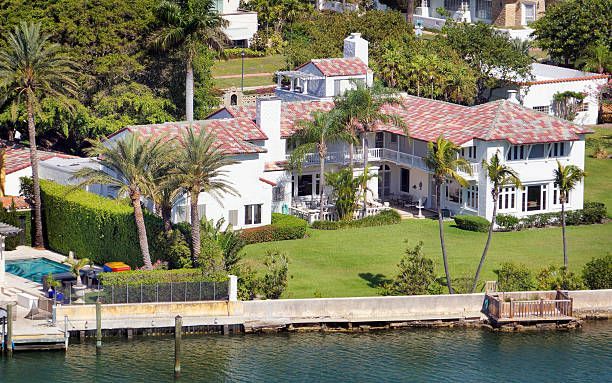 Large white house with red tile roof on waterfront, palm trees, green grass.