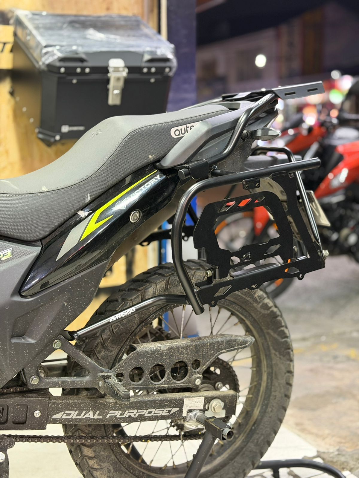 Motocicleta negra con portaequipajes laterales, caja de carga trasera y neumático de doble propósito.