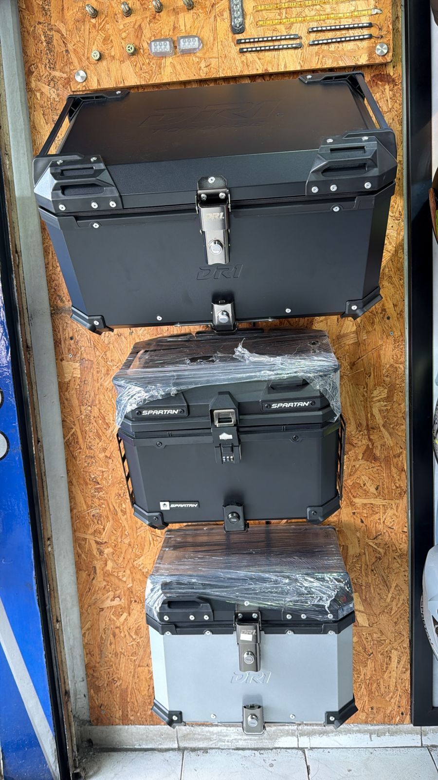 Tres baúles superiores de motocicleta negros y plateados exhibidos contra una pared de madera.