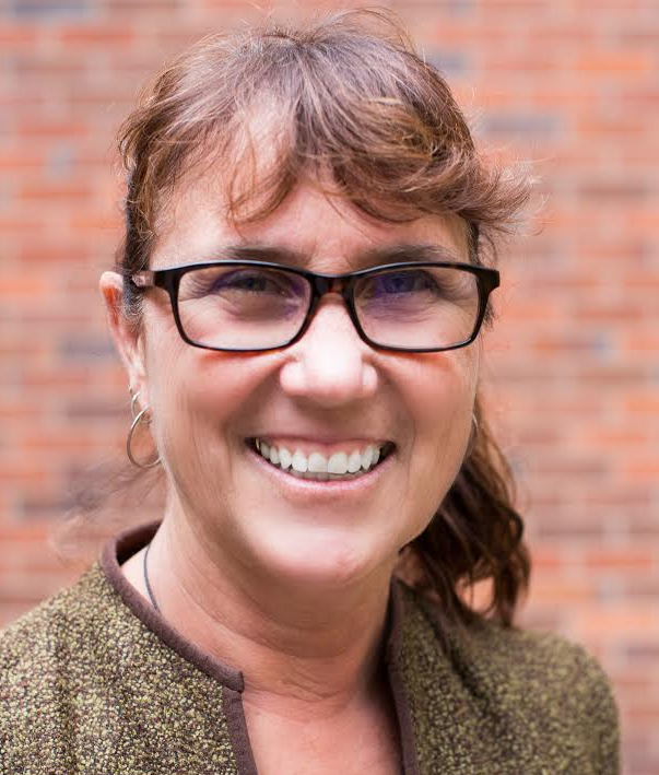a woman wearing glasses and a green jacket is smiling in front of a brick wall .