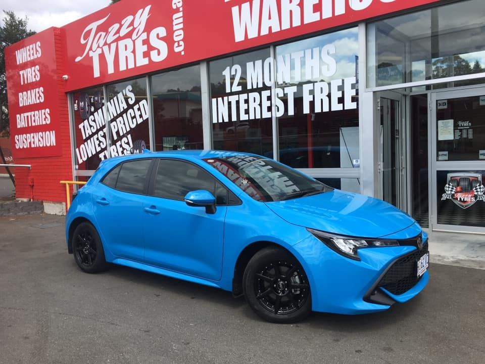 Blue Toyota Corolla in front of a tire store, black wheels, red building, sign.