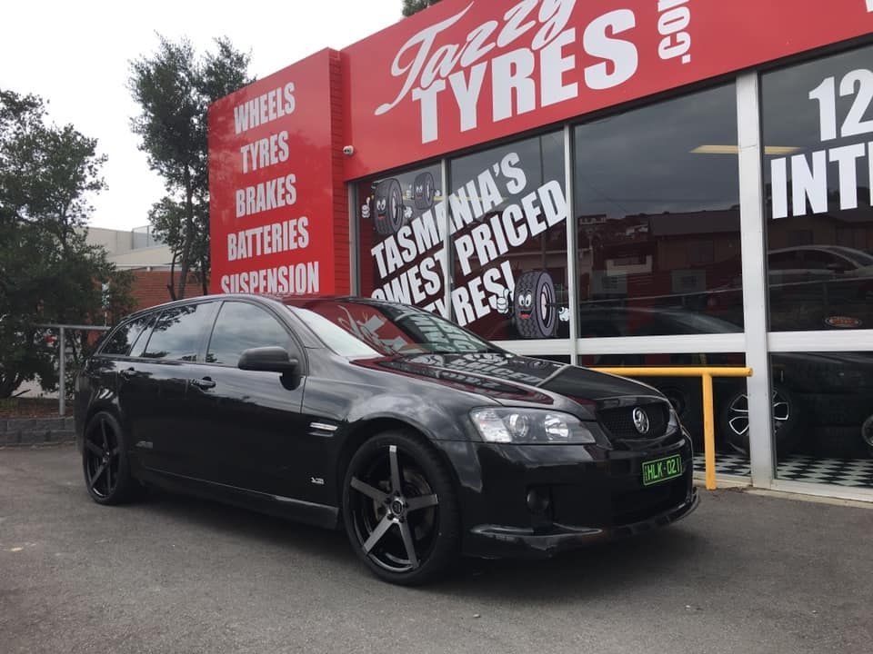 Black Holden wagon parked in front of Tazzy Tyres shop with red facade; advertisement for tires.