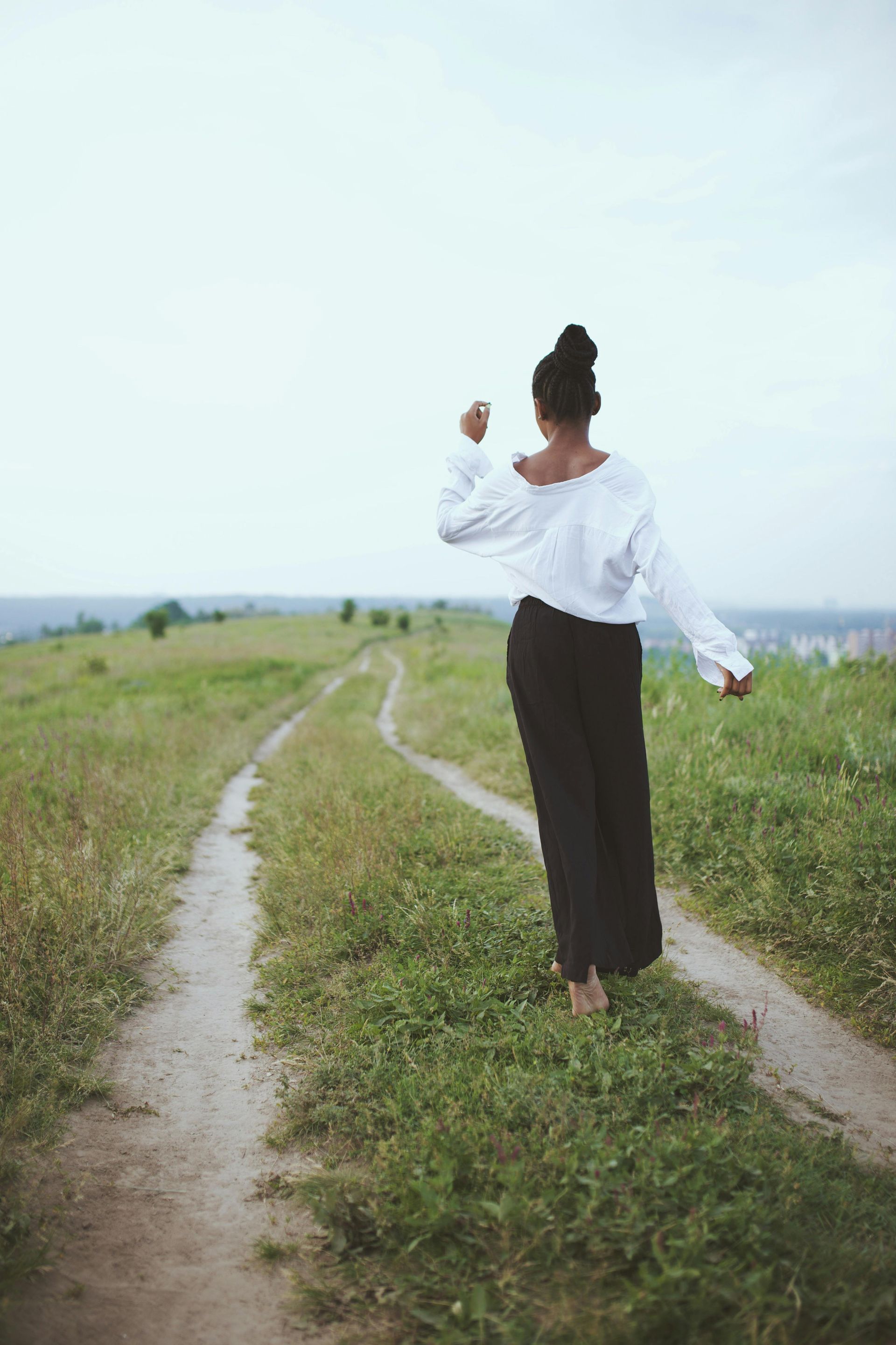Woman walking barefoot on a dirt path through a grassy field, wearing a white top and black pants.– holistic health