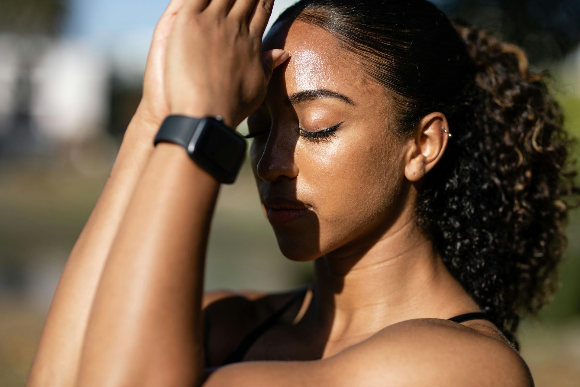 Woman with closed eyes, hands near forehead, sweating. Wearing smartwatch, outside.– mind-body connection