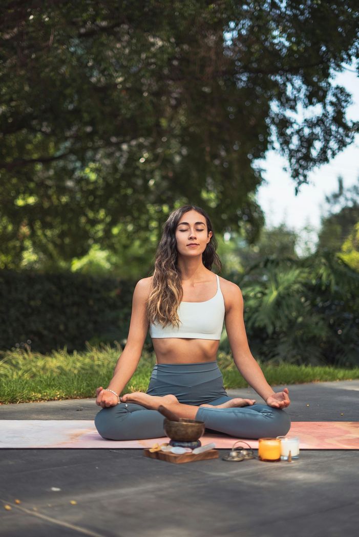 Woman in yoga pose, eyes closed, outdoors. Wearing white top, grey leggings. Meditation on a mat with objects.