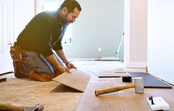 A man is kneeling down on the floor while installing a wooden floor.