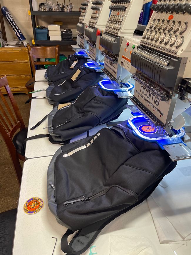 Multiple embroidery machines stitching logos onto dark backpacks lined up on a workspace table.