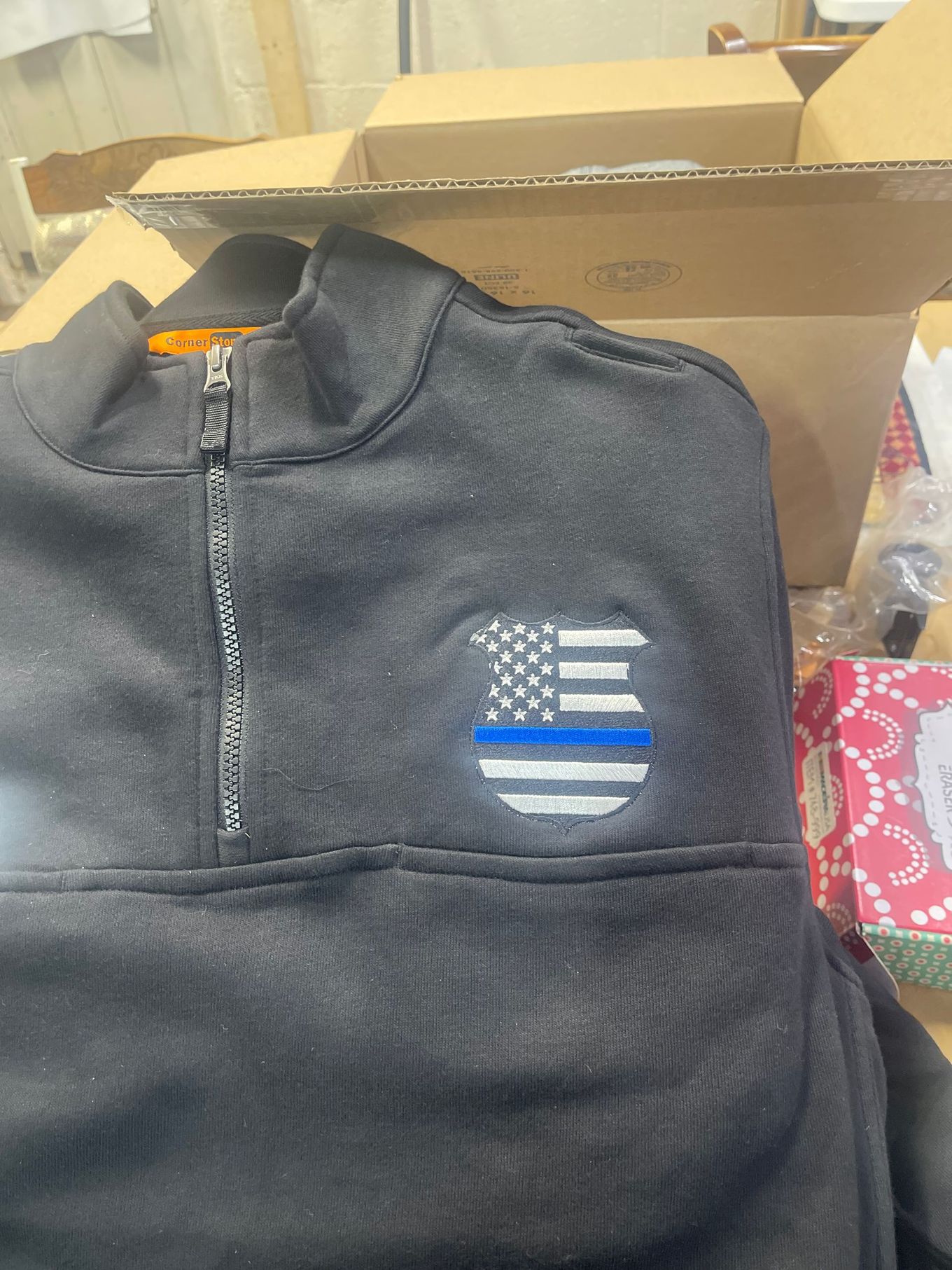 Black quarter-zip sweater featuring an embroidered shield-shaped patch of a flag with a thin blue horizontal stripe.