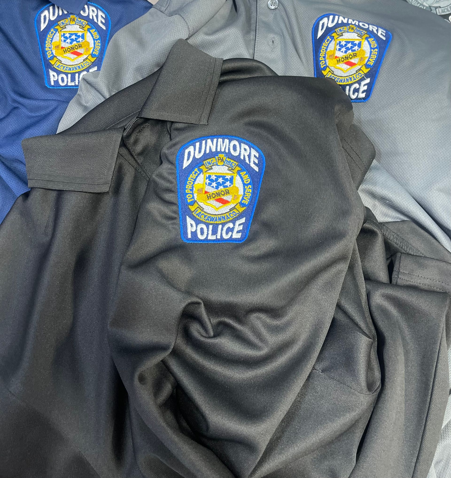 Three police shirts from the Dunmore Police Department, stacked in black, gray, and blue, each with a centered patch.