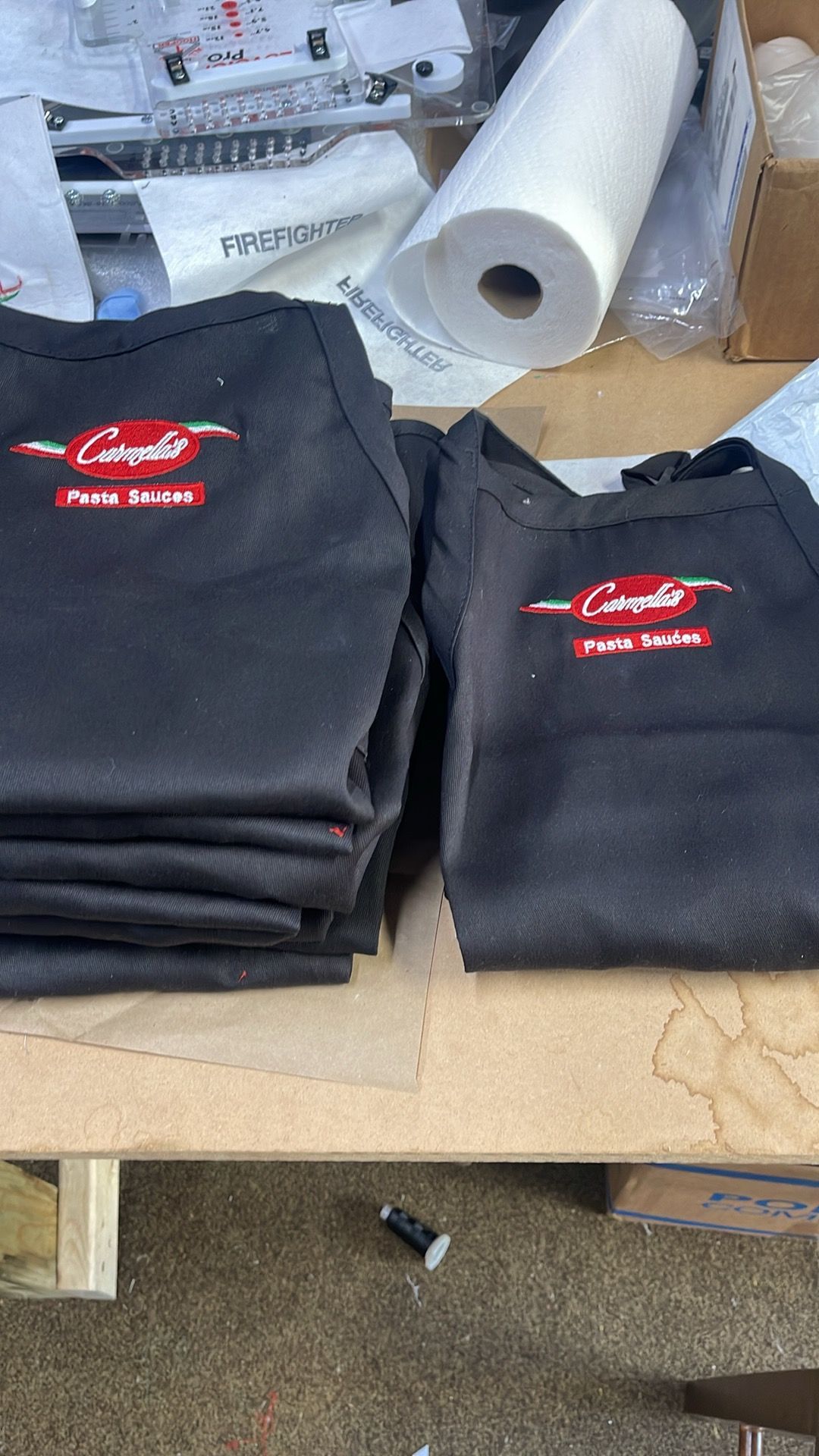 A stack of black aprons branded with the red and white Caterpillar logo sit on a workshop table near a roll of paper.