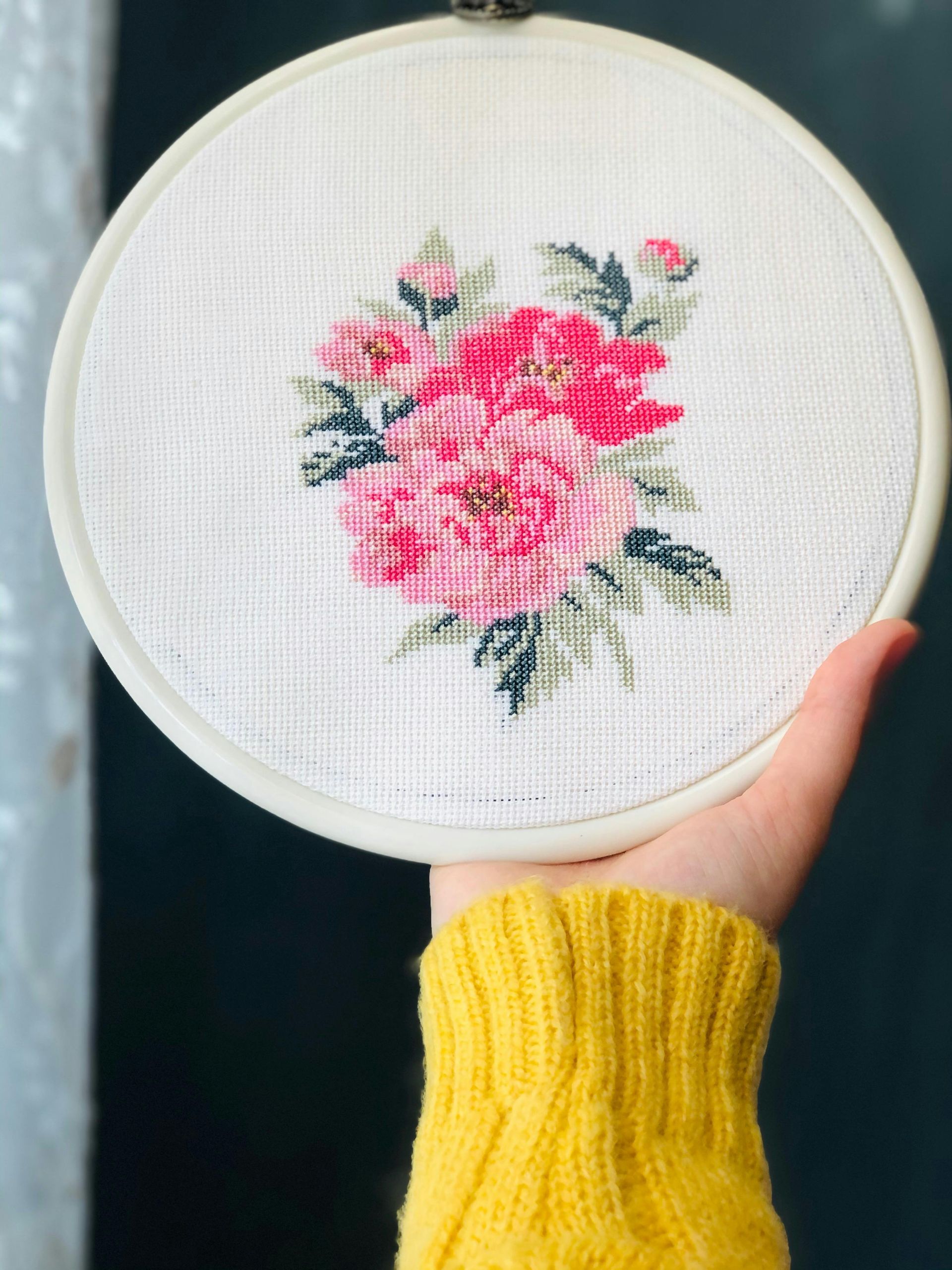 A hand in a yellow sweater holds a round embroidery hoop featuring a cross-stitched bouquet of pink flowers and green leaves.