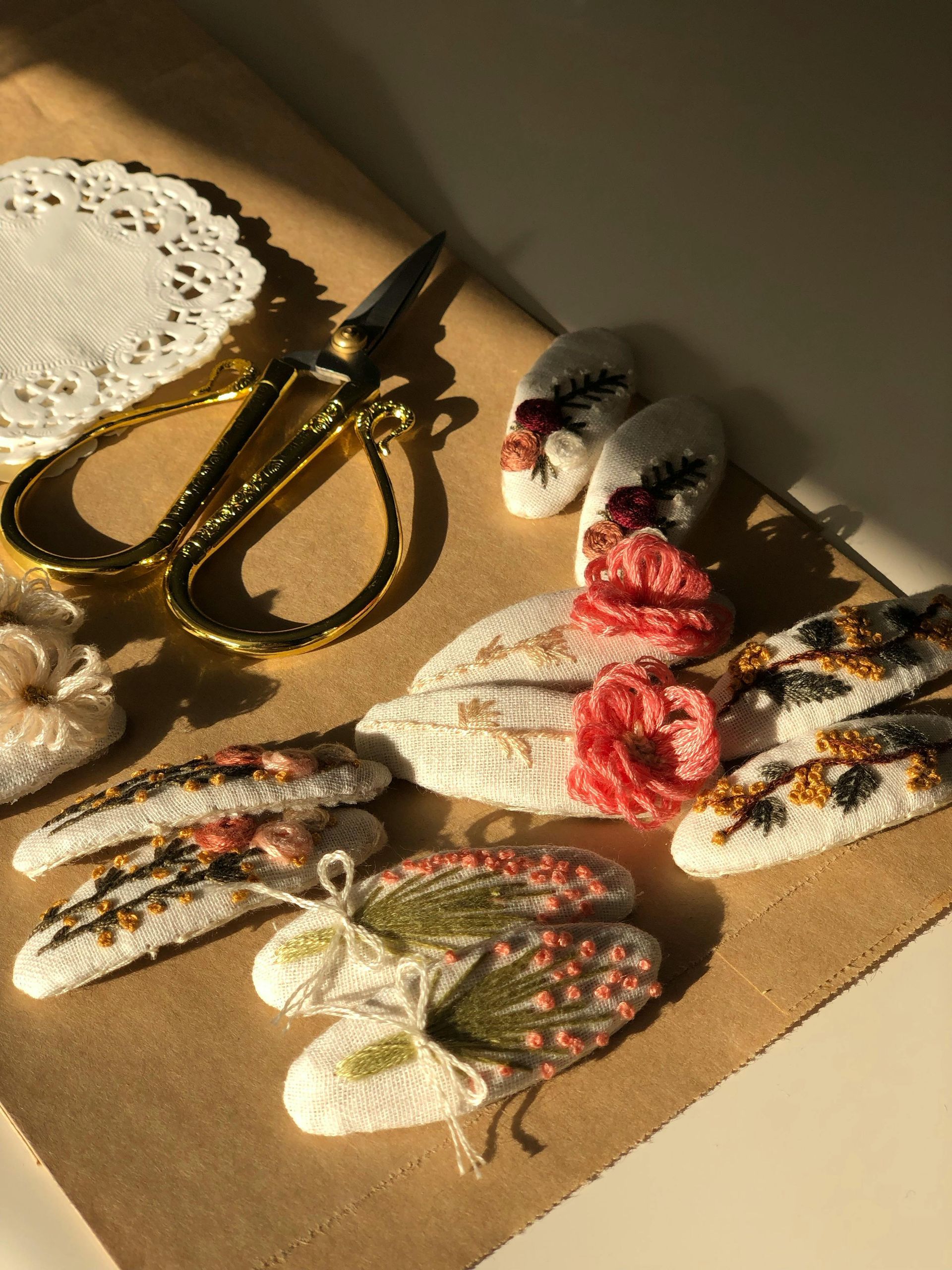 An assortment of handmade, embroidered hair clips with floral designs laid on brown paper next to gold scissors and a doily.