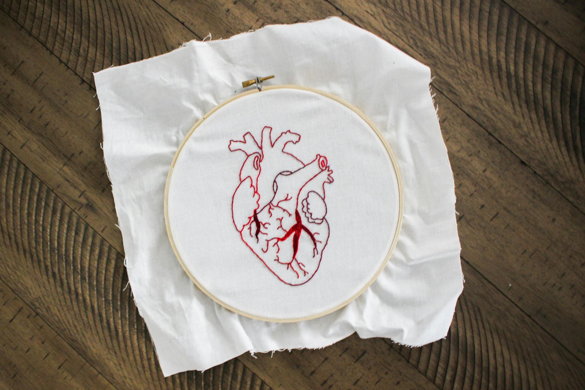 Red embroidery of an anatomical human heart in a circular wooden hoop, centered on a rustic wooden table.