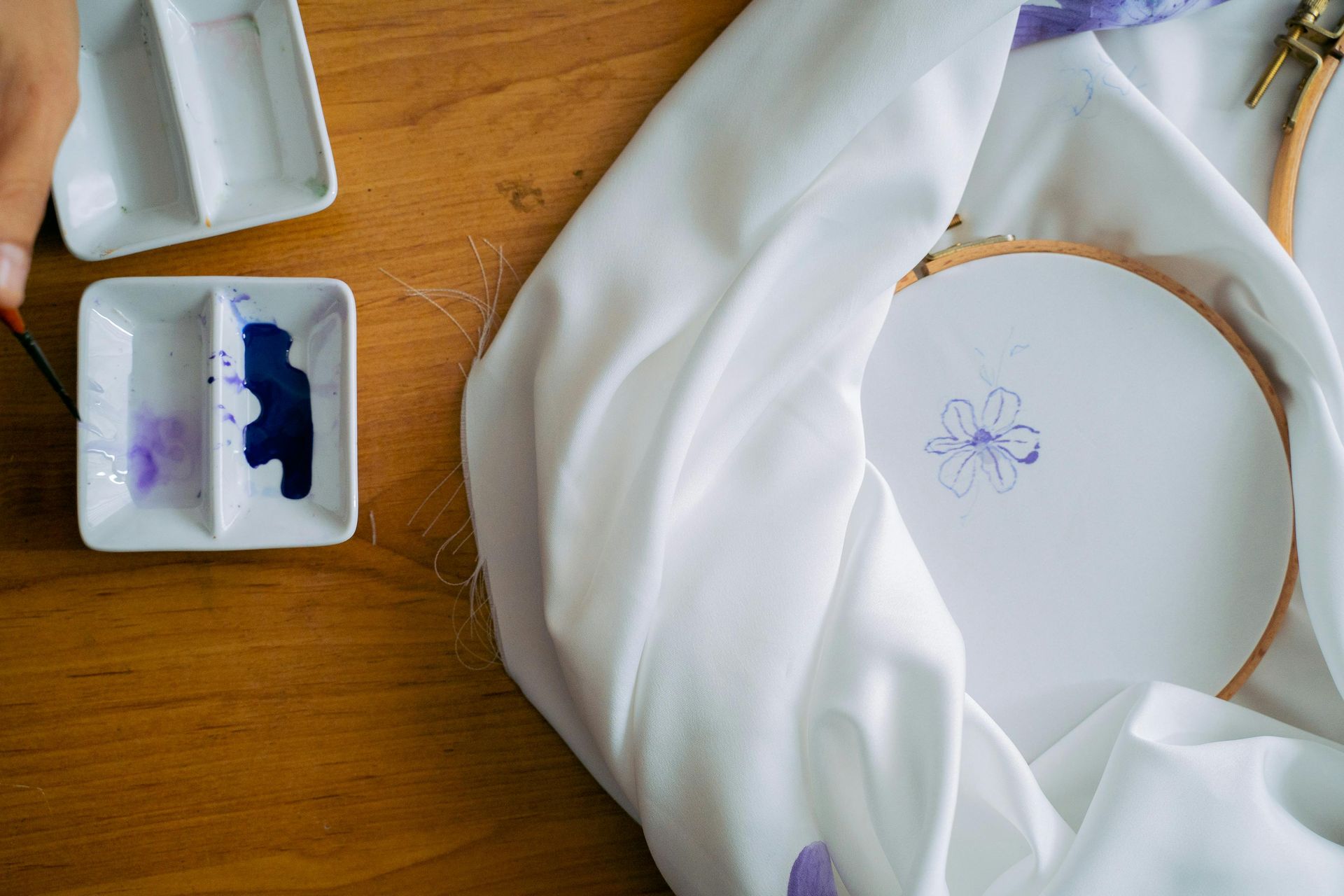 A hand paints a purple flower onto fabric held in a wooden embroidery hoop, next to a palette with purple and blue paint.