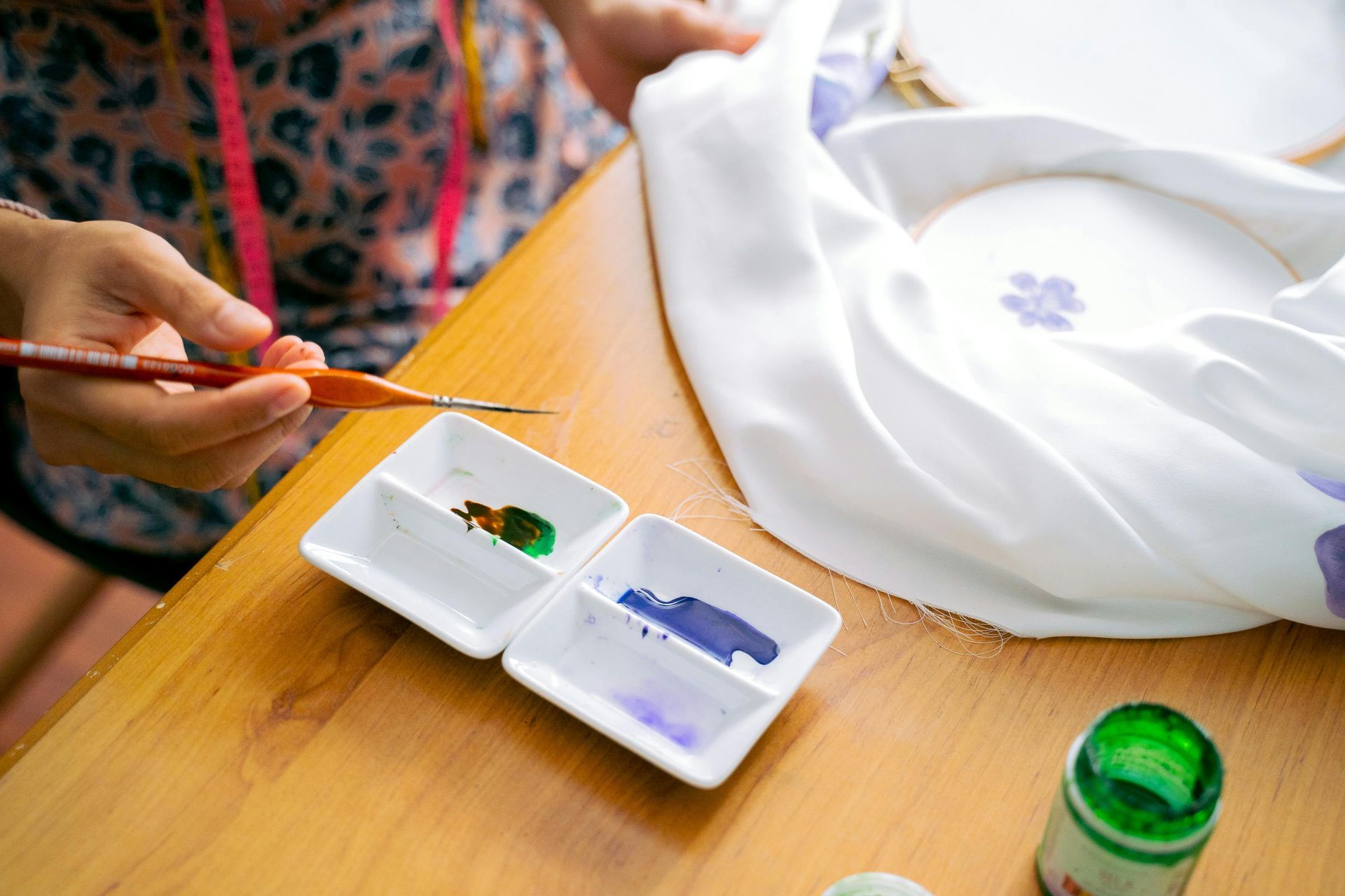 A person painting small purple flowers onto white fabric with a fine brush and a small white paint palette on a desk.