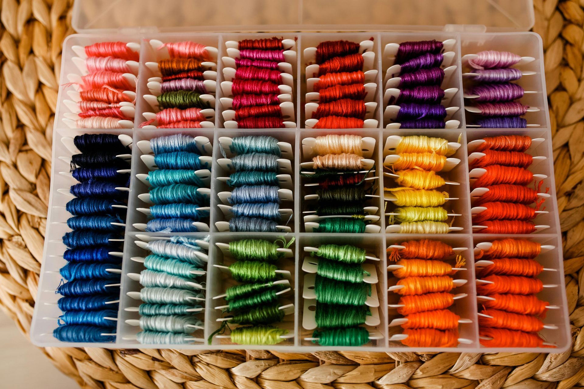 A clear plastic organizer box filled with colorful embroidery floss wound on cardboard bobbins, resting on a woven mat.