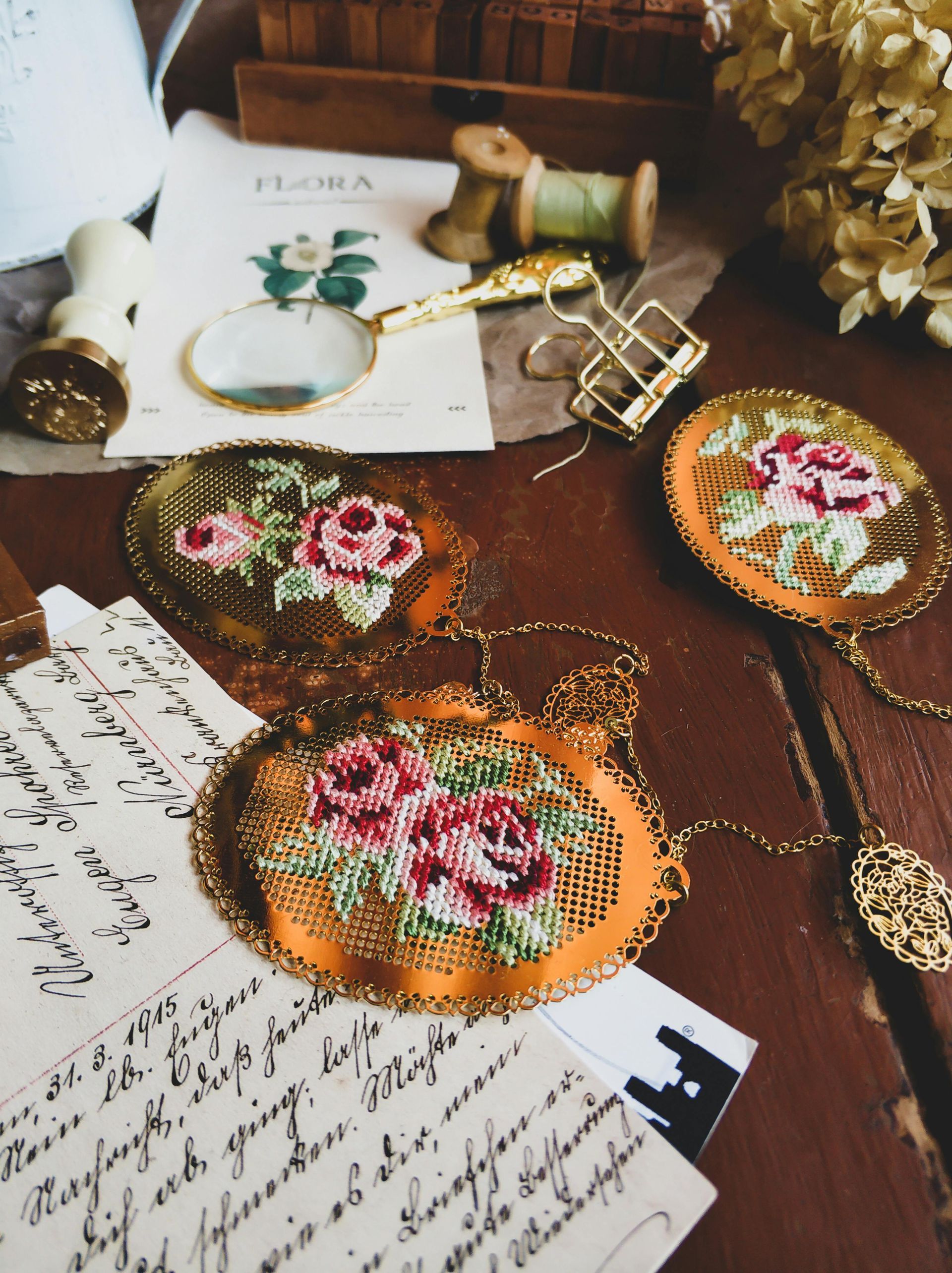 Three embroidered floral brooches rest on vintage handwritten letters and a desk with a magnifying glass and paper clips.
