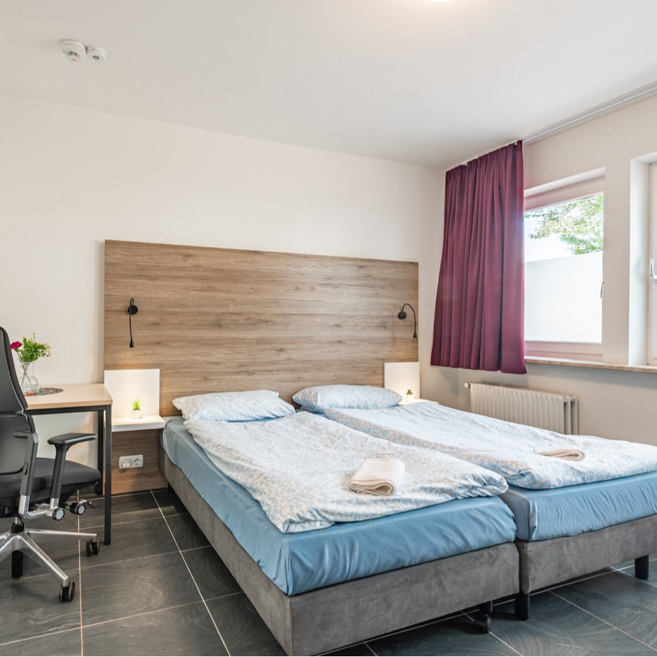 A hotel room with two beds, a desk, and a window with purple curtains.