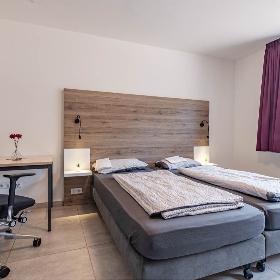 Bedroom with twin beds, wooden headboard with bedside lights, desk, and purple curtain.