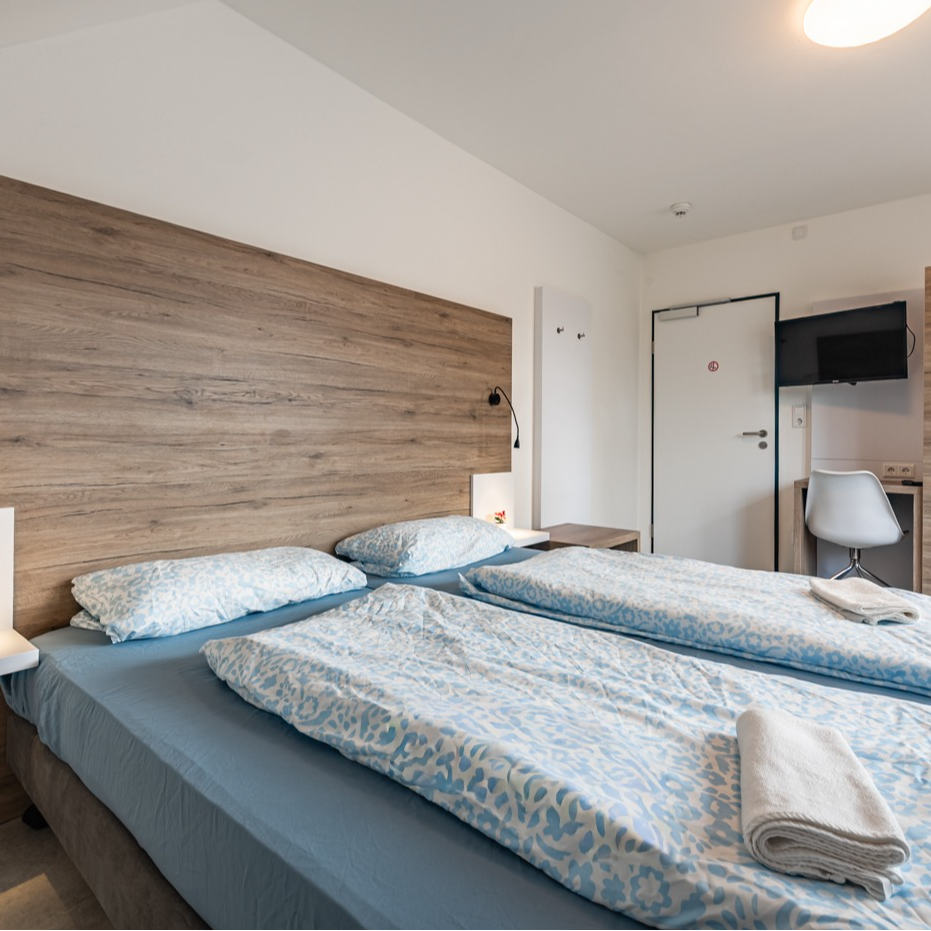 Bedroom with two beds, wood headboard, blue bedding, white walls, and a small desk.
