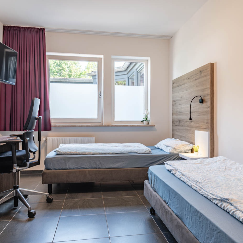 Bedroom with two beds, desk, windows, and dark curtain. Light-colored walls and dark tile flooring.