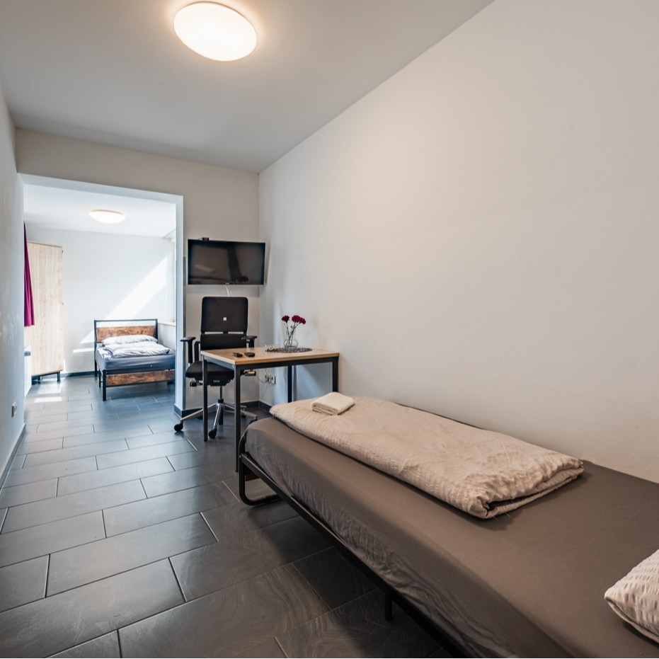 A simple, furnished room with a bed, desk, and TV. Another bed is visible in the adjacent room.