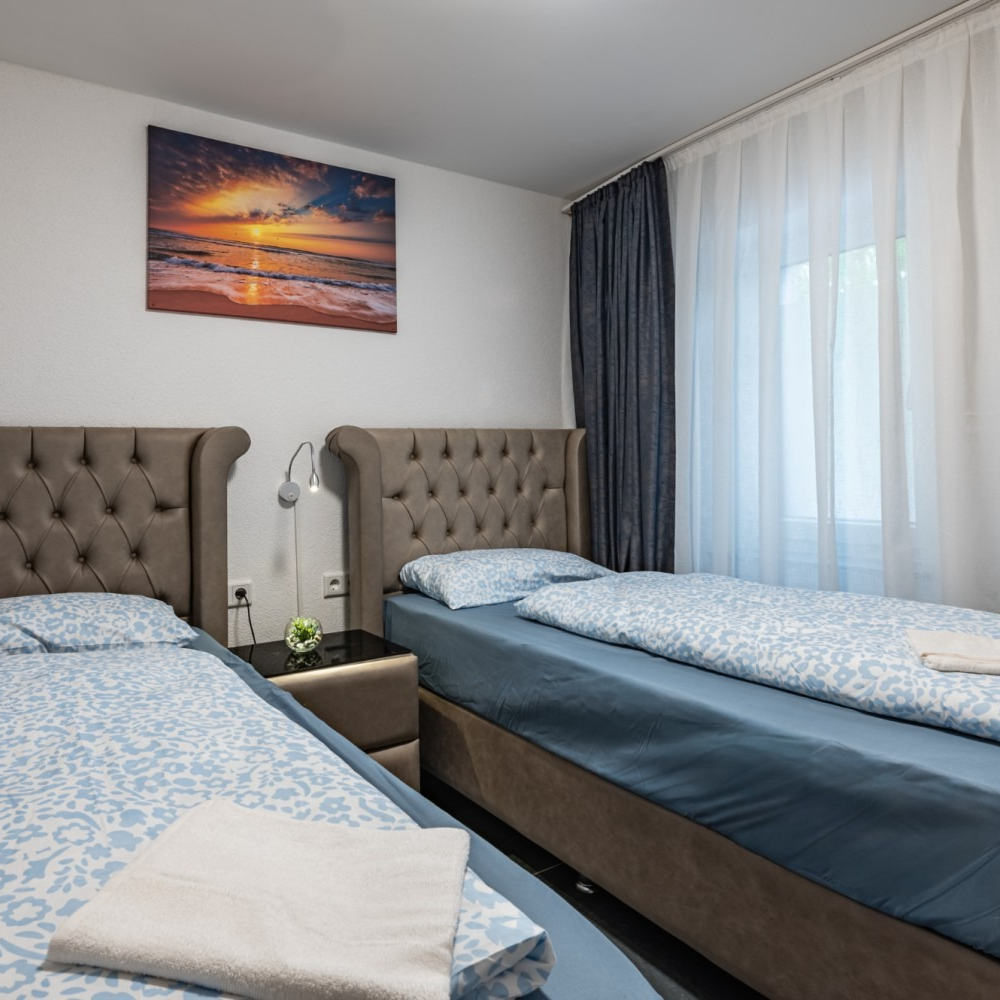 Two twin beds in a room, with a sunset art piece, and a window with dark blue curtains.