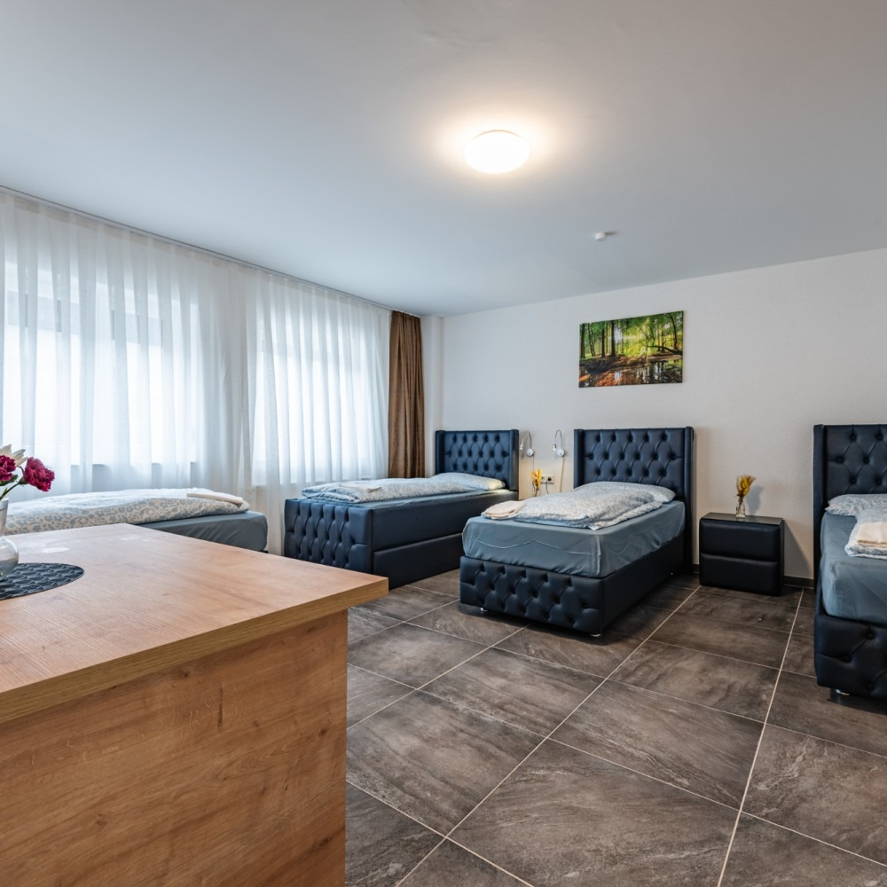 Four beds in a room, two by a window. Neutral-toned tile floor, wooden table in foreground. Green forest art on a wall.