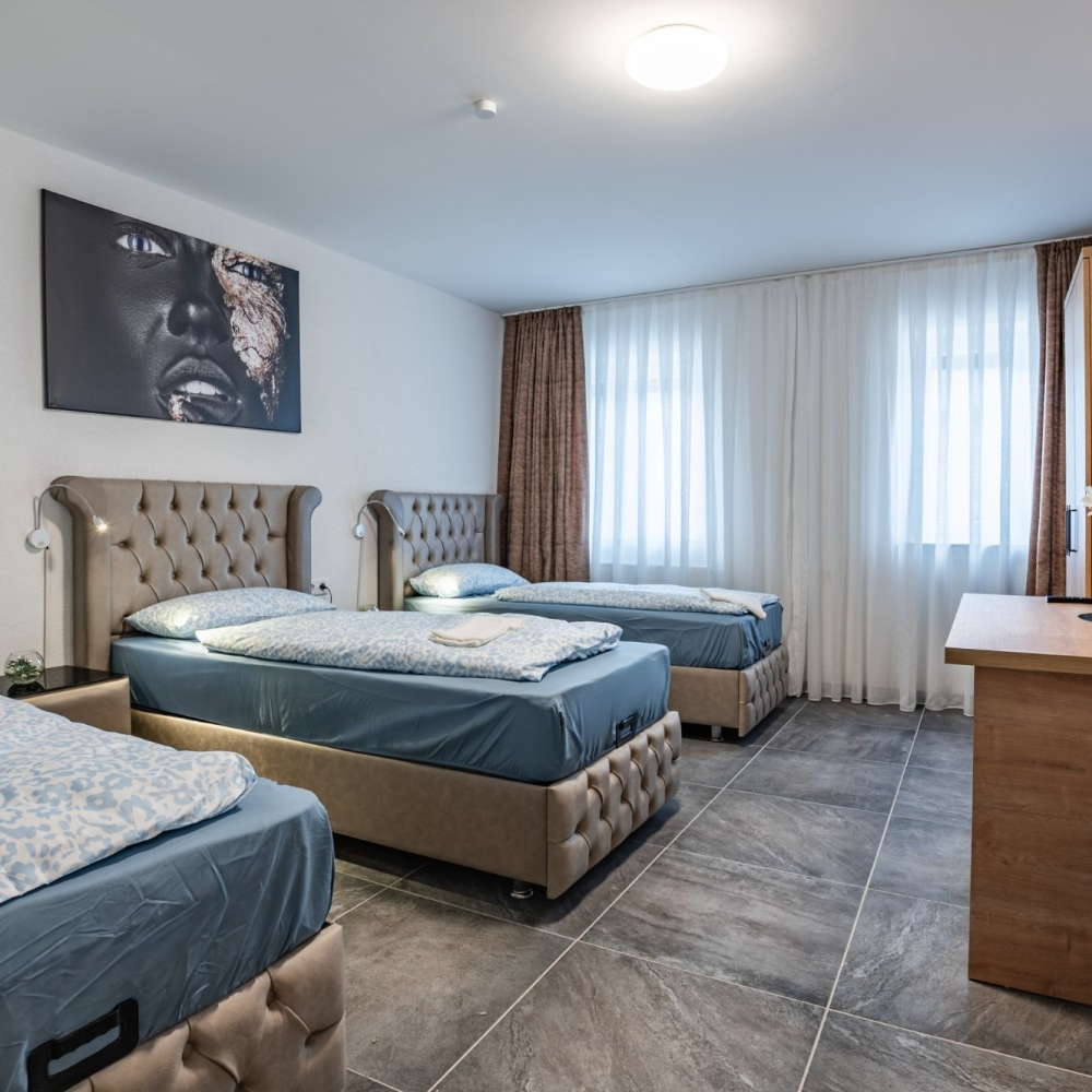 Bedroom with three twin beds, gray tiled floor, large artwork, and light filtering curtains.