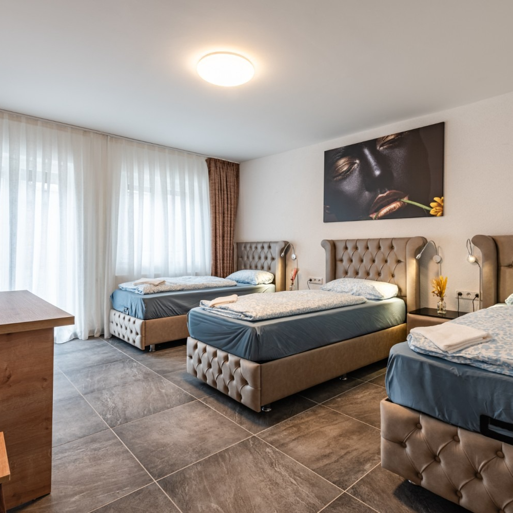 Three single beds in a room with neutral walls, large art piece, and windows with curtains.