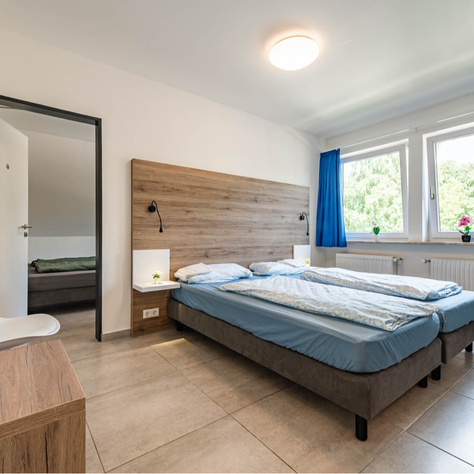 Bedroom with two twin beds, wooden headboard, large windows, and a doorway to another room.