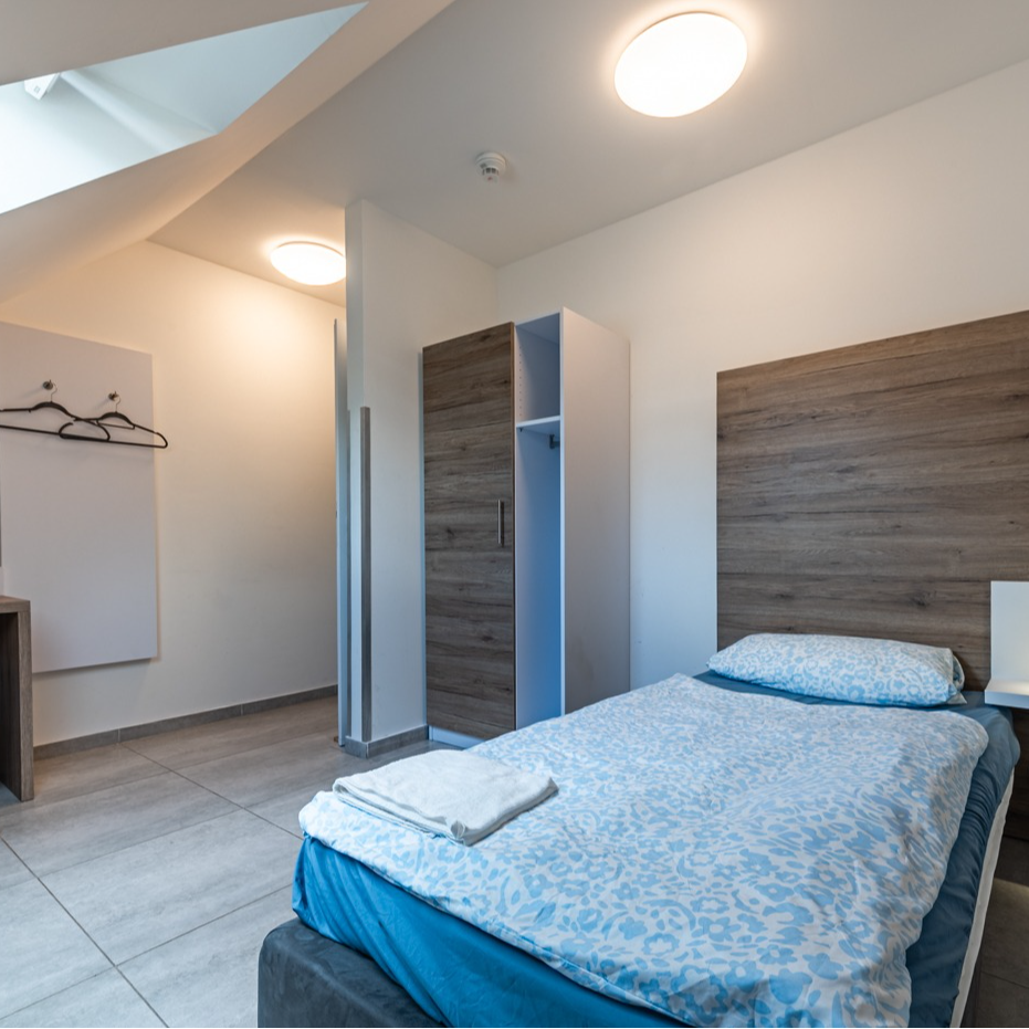 A minimalist hotel room with a single bed, wardrobe, and coat rack under a sloped ceiling.